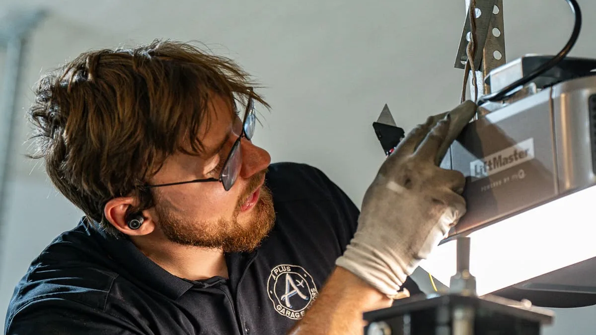 A technician in work gloves and safety glasses reaches up to a LiftMaster opener motor unit mounted on a garage ceiling.